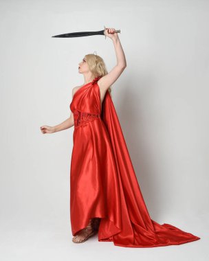 Eski mitolojik fantezi tanrıçası kılığında kırmızı ipek toga elbisesi ve taç giymiş güzel sarışın modelin tam boy portresi. Yürüyen bir poz, kılıç tutan bir silah, izole edilmiş bir stüdyo.