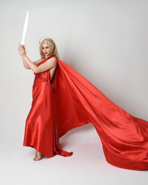 Eski mitolojik fantezi tanrıçası kılığında kırmızı ipek toga elbisesi ve taç giymiş güzel sarışın modelin tam boy portresi. Yürüyen bir poz, kılıç tutan bir silah, izole edilmiş bir stüdyo.