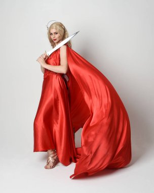 Eski mitolojik fantezi tanrıçası kılığında kırmızı ipek toga elbisesi ve taç giymiş güzel sarışın modelin tam boy portresi. Yürüyen bir poz, kılıç tutan bir silah, izole edilmiş bir stüdyo.