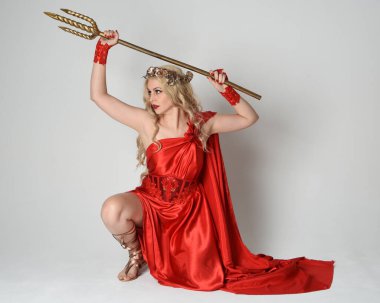 Full length portrait of beautiful blonde model dressed as ancient mythological fantasy goddess in flowing red silk toga gown, crown. kneeling pose,  golden trident weapon, isolated studio background clipart
