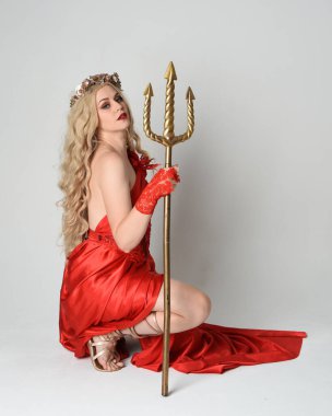 Eski mitolojik fantezi tanrıçası kılığında kırmızı ipek toga elbisesi ve taç giymiş güzel sarışın modelin tam boy portresi. Diz çökme pozisyonu, altın mızrak, izole edilmiş stüdyo arka planı.