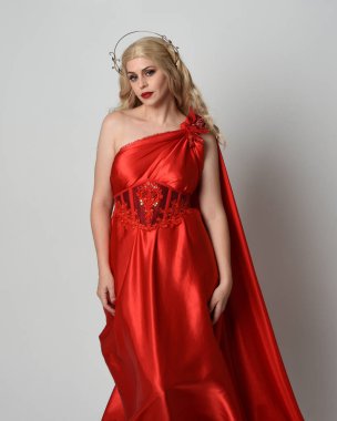 Kırmızı ipek toga elbisesi ve taç giyen, antik mitolojik fantezi tanrıçası gibi giyinmiş güzel sarışın mankenin yakın portresi. Stüdyoda izole edilmiş zarif bir poz..