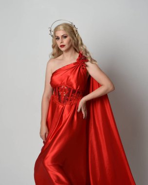 Kırmızı ipek toga elbisesi ve taç giyen, antik mitolojik fantezi tanrıçası gibi giyinmiş güzel sarışın mankenin yakın portresi. Stüdyoda izole edilmiş zarif bir poz..