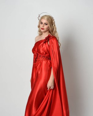 Kırmızı ipek toga elbisesi ve taç giyen, antik mitolojik fantezi tanrıçası gibi giyinmiş güzel sarışın mankenin yakın portresi. Stüdyoda izole edilmiş zarif bir poz..