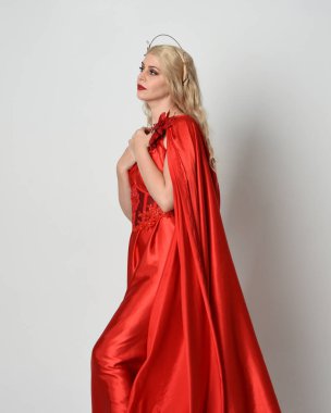 Kırmızı ipek toga elbisesi ve taç giyen, antik mitolojik fantezi tanrıçası gibi giyinmiş güzel sarışın mankenin yakın portresi. Stüdyoda izole edilmiş zarif bir poz..