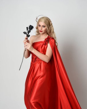 Kırmızı ipek toga elbisesi ve taç giyen, antik mitolojik fantezi tanrıçası gibi giyinmiş güzel sarışın mankenin yakın portresi. Gül çiçeği taşıyan stüdyo arka planı.