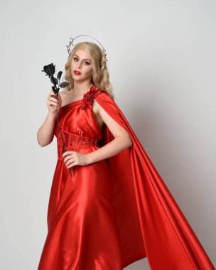 Kırmızı ipek toga elbisesi ve taç giyen, antik mitolojik fantezi tanrıçası gibi giyinmiş güzel sarışın mankenin yakın portresi. Gül çiçeği taşıyan stüdyo arka planı.