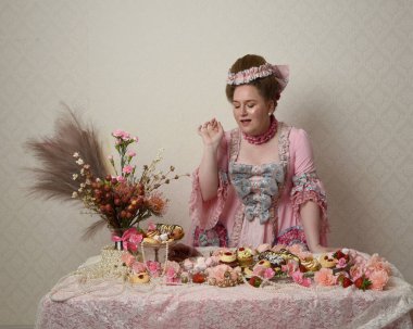 Zengin pembe bir elbise giyen sevimli bir kadın modelin portresi. Tarihi bir Fransız barok asilzadesinin kostümü. Hamur işleri, çiçekler ve zengin mücevherlerle dolu bir masada, hoş bir ziyafette yemek ve tatlı yemek yemek..