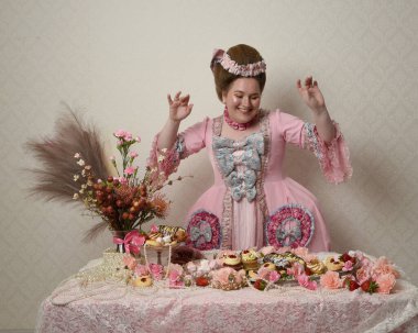 Zengin pembe bir elbise giyen sevimli bir kadın modelin portresi. Tarihi bir Fransız barok asilzadesinin kostümü. Hamur işleri, çiçekler ve zengin mücevherlerle dolu bir masada, hoş bir ziyafette yemek ve tatlı yemek yemek..