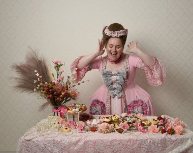 Zengin pembe bir elbise giyen sevimli bir kadın modelin portresi. Tarihi bir Fransız barok asilzadesinin kostümü. Hamur işleri, çiçekler ve zengin mücevherlerle dolu bir masada, hoş bir ziyafette yemek ve tatlı yemek yemek..