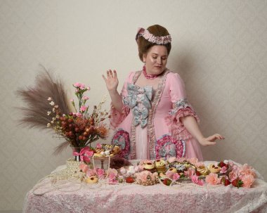 Zengin pembe bir elbise giyen sevimli bir kadın modelin portresi. Tarihi bir Fransız barok asilzadesinin kostümü. Hamur işleri, çiçekler ve zengin mücevherlerle dolu bir masada, hoş bir ziyafette yemek ve tatlı yemek yemek..