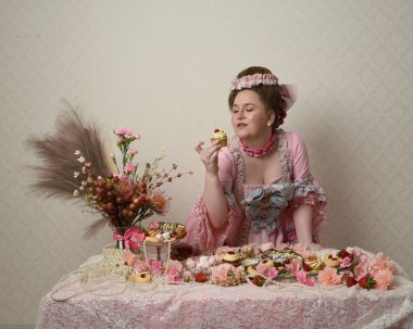 Zengin pembe bir elbise giyen sevimli bir kadın modelin portresi. Tarihi bir Fransız barok asilzadesinin kostümü. Hamur işleri, çiçekler ve zengin mücevherlerle dolu bir masada, hoş bir ziyafette yemek ve tatlı yemek yemek..