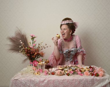 Zengin pembe bir elbise giyen sevimli bir kadın modelin portresi. Tarihi bir Fransız barok asilzadesinin kostümü. Hamur işleri, çiçekler ve zengin mücevherlerle dolu bir masada, hoş bir ziyafette yemek ve tatlı yemek yemek..