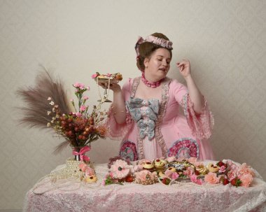 Zengin pembe bir elbise giyen sevimli bir kadın modelin portresi. Tarihi bir Fransız barok asilzadesinin kostümü. Hamur işleri, çiçekler ve zengin mücevherlerle dolu bir masada, hoş bir ziyafette yemek ve tatlı yemek yemek..