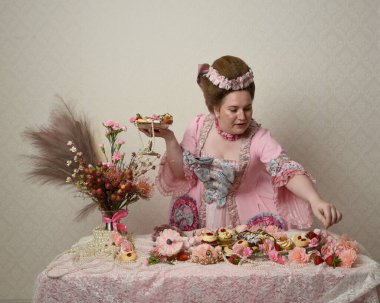 Zengin pembe bir elbise giyen sevimli bir kadın modelin portresi. Tarihi bir Fransız barok asilzadesinin kostümü. Hamur işleri, çiçekler ve zengin mücevherlerle dolu bir masada, hoş bir ziyafette yemek ve tatlı yemek yemek..