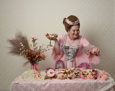 Zengin pembe bir elbise giyen sevimli bir kadın modelin portresi. Tarihi bir Fransız barok asilzadesinin kostümü. Hamur işleri, çiçekler ve zengin mücevherlerle dolu bir masada, hoş bir ziyafette yemek ve tatlı yemek yemek..