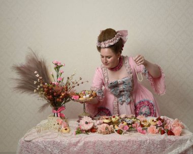 Zengin pembe bir elbise giyen sevimli bir kadın modelin portresi. Tarihi bir Fransız barok asilzadesinin kostümü. Hamur işleri, çiçekler ve zengin mücevherlerle dolu bir masada, hoş bir ziyafette yemek ve tatlı yemek yemek..