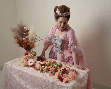 Zengin pembe bir elbise giyen sevimli bir kadın modelin portresi. Tarihi bir Fransız barok asilzadesinin kostümü. Hamur işleri, çiçekler ve zengin mücevherlerle dolu bir masada, hoş bir ziyafette yemek ve tatlı yemek yemek..