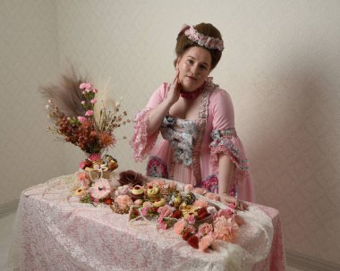 Zengin pembe bir elbise giyen sevimli bir kadın modelin portresi. Tarihi bir Fransız barok asilzadesinin kostümü. Hamur işleri, çiçekler ve zengin mücevherlerle dolu bir masada, hoş bir ziyafette yemek ve tatlı yemek yemek..