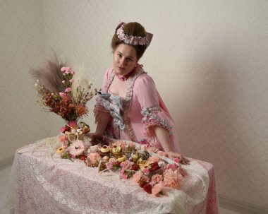 Zengin pembe bir elbise giyen sevimli bir kadın modelin portresi. Tarihi bir Fransız barok asilzadesinin kostümü. Hamur işleri, çiçekler ve zengin mücevherlerle dolu bir masada, hoş bir ziyafette yemek ve tatlı yemek yemek..