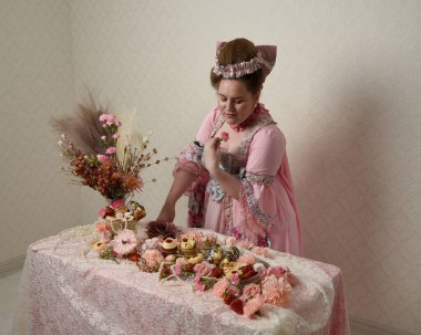 Zengin pembe bir elbise giyen sevimli bir kadın modelin portresi. Tarihi bir Fransız barok asilzadesinin kostümü. Hamur işleri, çiçekler ve zengin mücevherlerle dolu bir masada, hoş bir ziyafette yemek ve tatlı yemek yemek..