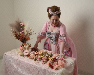 Zengin pembe bir elbise giyen sevimli bir kadın modelin portresi. Tarihi bir Fransız barok asilzadesinin kostümü. Hamur işleri, çiçekler ve zengin mücevherlerle dolu bir masada, hoş bir ziyafette yemek ve tatlı yemek yemek..