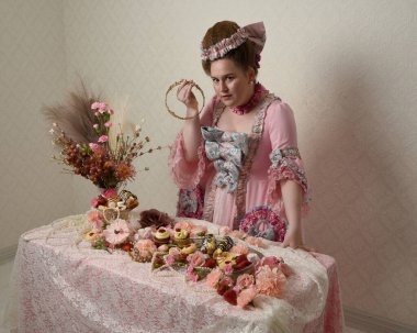 Zengin pembe bir elbise giyen sevimli bir kadın modelin portresi. Tarihi bir Fransız barok asilzadesinin kostümü. Hamur işleri, çiçekler ve zengin mücevherlerle dolu bir masada, hoş bir ziyafette yemek ve tatlı yemek yemek..