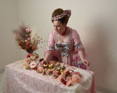 Zengin pembe bir elbise giyen sevimli bir kadın modelin portresi. Tarihi bir Fransız barok asilzadesinin kostümü. Hamur işleri, çiçekler ve zengin mücevherlerle dolu bir masada, hoş bir ziyafette yemek ve tatlı yemek yemek..