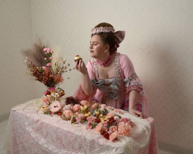 Zengin pembe bir elbise giyen sevimli bir kadın modelin portresi. Tarihi bir Fransız barok asilzadesinin kostümü. Hamur işleri, çiçekler ve zengin mücevherlerle dolu bir masada, hoş bir ziyafette yemek ve tatlı yemek yemek..