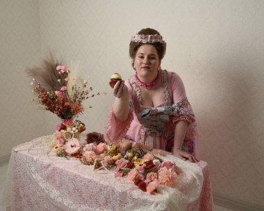 Zengin pembe bir elbise giyen sevimli bir kadın modelin portresi. Tarihi bir Fransız barok asilzadesinin kostümü. Hamur işleri, çiçekler ve zengin mücevherlerle dolu bir masada, hoş bir ziyafette yemek ve tatlı yemek yemek..