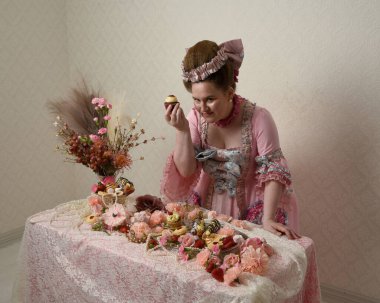 Zengin pembe bir elbise giyen sevimli bir kadın modelin portresi. Tarihi bir Fransız barok asilzadesinin kostümü. Hamur işleri, çiçekler ve zengin mücevherlerle dolu bir masada, hoş bir ziyafette yemek ve tatlı yemek yemek..