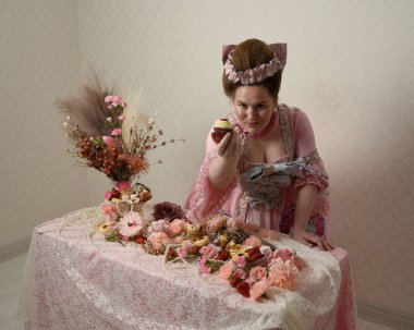 Zengin pembe bir elbise giyen sevimli bir kadın modelin portresi. Tarihi bir Fransız barok asilzadesinin kostümü. Hamur işleri, çiçekler ve zengin mücevherlerle dolu bir masada, hoş bir ziyafette yemek ve tatlı yemek yemek..