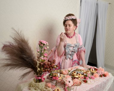 Zengin pembe bir elbise giyen sevimli bir kadın modelin portresi. Tarihi bir Fransız barok asilzadesinin kostümü. Hamur işleri, çiçekler ve zengin mücevherlerle dolu bir masada, hoş bir ziyafette yemek ve tatlı yemek yemek..
