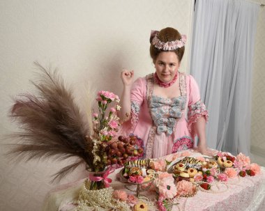 Zengin pembe bir elbise giyen sevimli bir kadın modelin portresi. Tarihi bir Fransız barok asilzadesinin kostümü. Hamur işleri, çiçekler ve zengin mücevherlerle dolu bir masada, hoş bir ziyafette yemek ve tatlı yemek yemek..