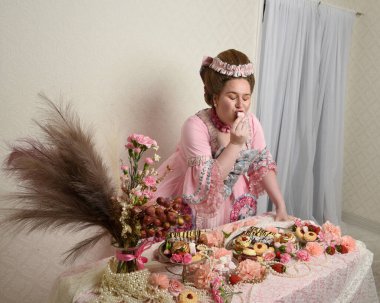 Zengin pembe bir elbise giyen sevimli bir kadın modelin portresi. Tarihi bir Fransız barok asilzadesinin kostümü. Hamur işleri, çiçekler ve zengin mücevherlerle dolu bir masada, hoş bir ziyafette yemek ve tatlı yemek yemek..