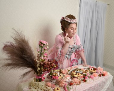 Zengin pembe bir elbise giyen sevimli bir kadın modelin portresi. Tarihi bir Fransız barok asilzadesinin kostümü. Hamur işleri, çiçekler ve zengin mücevherlerle dolu bir masada, hoş bir ziyafette yemek ve tatlı yemek yemek..