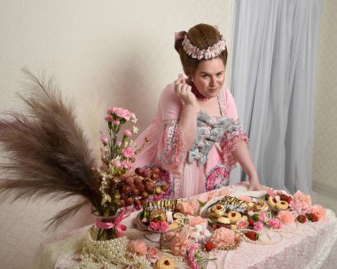 Zengin pembe bir elbise giyen sevimli bir kadın modelin portresi. Tarihi bir Fransız barok asilzadesinin kostümü. Hamur işleri, çiçekler ve zengin mücevherlerle dolu bir masada, hoş bir ziyafette yemek ve tatlı yemek yemek..