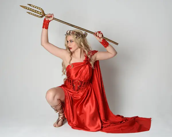 stock image Full length portrait of beautiful blonde model dressed as ancient mythological fantasy goddess in flowing red silk toga gown, crown. kneeling pose,  golden trident weapon, isolated studio background