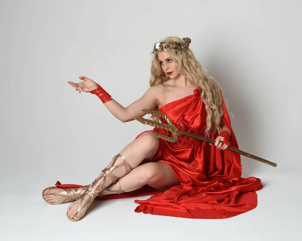 stock image Full length portrait of beautiful blonde model dressed as ancient mythological fantasy goddess in flowing red silk toga gown, crown. kneeling pose,  golden trident weapon, isolated studio background