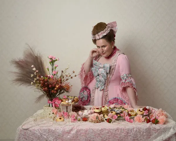 Zengin pembe bir elbise giyen sevimli bir kadın modelin portresi. Tarihi bir Fransız barok asilzadesinin kostümü. Hamur işleri, çiçekler ve zengin mücevherlerle dolu bir masada, hoş bir ziyafette yemek ve tatlı yemek yemek..