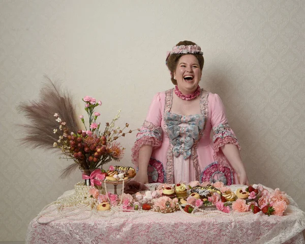 Zengin pembe bir elbise giyen sevimli bir kadın modelin portresi. Tarihi bir Fransız barok asilzadesinin kostümü. Hamur işleri, çiçekler ve zengin mücevherlerle dolu bir masada, hoş bir ziyafette yemek ve tatlı yemek yemek..