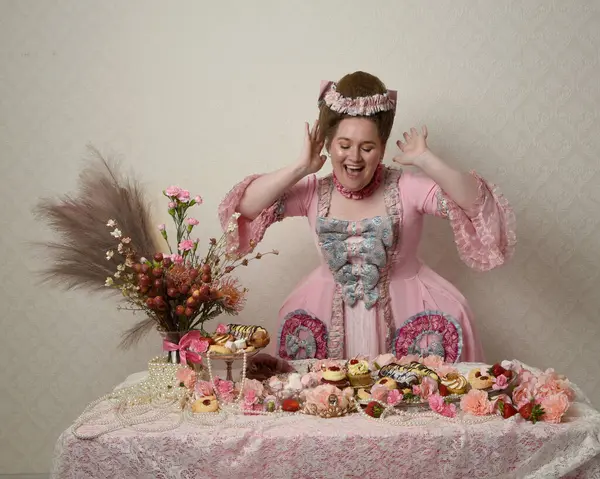 Zengin pembe bir elbise giyen sevimli bir kadın modelin portresi. Tarihi bir Fransız barok asilzadesinin kostümü. Hamur işleri, çiçekler ve zengin mücevherlerle dolu bir masada, hoş bir ziyafette yemek ve tatlı yemek yemek..