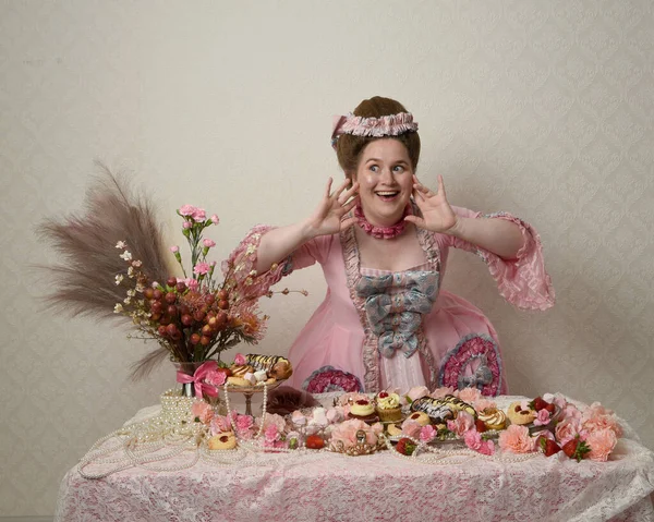 Zengin pembe bir elbise giyen sevimli bir kadın modelin portresi. Tarihi bir Fransız barok asilzadesinin kostümü. Hamur işleri, çiçekler ve zengin mücevherlerle dolu bir masada, hoş bir ziyafette yemek ve tatlı yemek yemek..