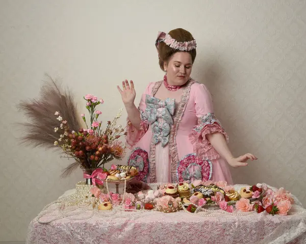 Zengin pembe bir elbise giyen sevimli bir kadın modelin portresi. Tarihi bir Fransız barok asilzadesinin kostümü. Hamur işleri, çiçekler ve zengin mücevherlerle dolu bir masada, hoş bir ziyafette yemek ve tatlı yemek yemek..