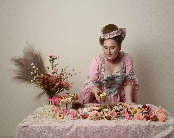 Zengin pembe bir elbise giyen sevimli bir kadın modelin portresi. Tarihi bir Fransız barok asilzadesinin kostümü. Hamur işleri, çiçekler ve zengin mücevherlerle dolu bir masada, hoş bir ziyafette yemek ve tatlı yemek yemek..