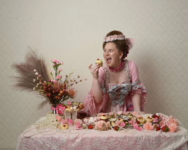 Zengin pembe bir elbise giyen sevimli bir kadın modelin portresi. Tarihi bir Fransız barok asilzadesinin kostümü. Hamur işleri, çiçekler ve zengin mücevherlerle dolu bir masada, hoş bir ziyafette yemek ve tatlı yemek yemek..