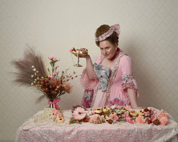 Zengin pembe bir elbise giyen sevimli bir kadın modelin portresi. Tarihi bir Fransız barok asilzadesinin kostümü. Hamur işleri, çiçekler ve zengin mücevherlerle dolu bir masada, hoş bir ziyafette yemek ve tatlı yemek yemek..
