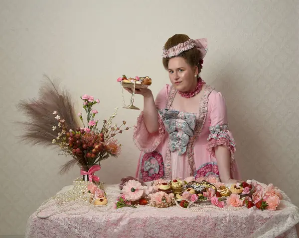 Zengin pembe bir elbise giyen sevimli bir kadın modelin portresi. Tarihi bir Fransız barok asilzadesinin kostümü. Hamur işleri, çiçekler ve zengin mücevherlerle dolu bir masada, hoş bir ziyafette yemek ve tatlı yemek yemek..