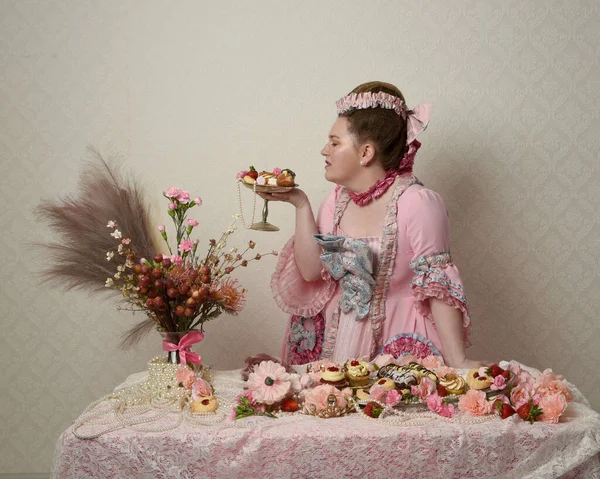 Zengin pembe bir elbise giyen sevimli bir kadın modelin portresi. Tarihi bir Fransız barok asilzadesinin kostümü. Hamur işleri, çiçekler ve zengin mücevherlerle dolu bir masada, hoş bir ziyafette yemek ve tatlı yemek yemek..