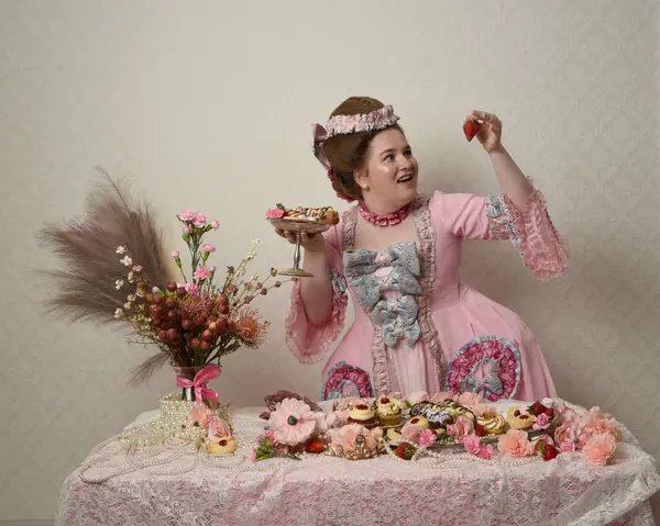 Zengin pembe bir elbise giyen sevimli bir kadın modelin portresi. Tarihi bir Fransız barok asilzadesinin kostümü. Hamur işleri, çiçekler ve zengin mücevherlerle dolu bir masada, hoş bir ziyafette yemek ve tatlı yemek yemek..